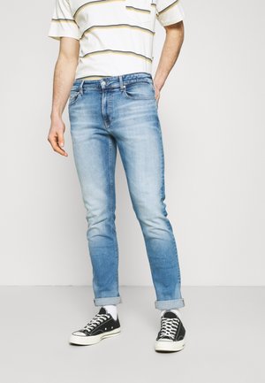 Homme portant un jean skinny bleu clair retroussé, des chaussettes blanches, des baskets noires et une chemise blanche à manches courtes rayée.