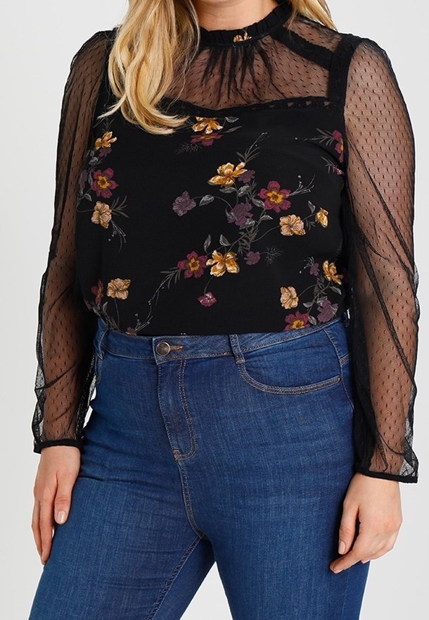 Femme portant un chemisier long noir transparent à manches longues avec des motifs floraux et un jean bleu taille haute sur un fond uni.