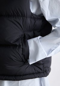 Svart puffervest med en jevn tekstur, som har en lomme foran. Kombinert med en lyseblå skjorte, som viser kontrasterende materialer.