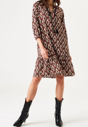 Frau trägt ein knielanges, gemustertes Kleid mit Dreiviertelärmeln und schwarze, spitz zulaufende Stiefel bis zur Mitte der Wade, steht mit gespreizten Beinen.