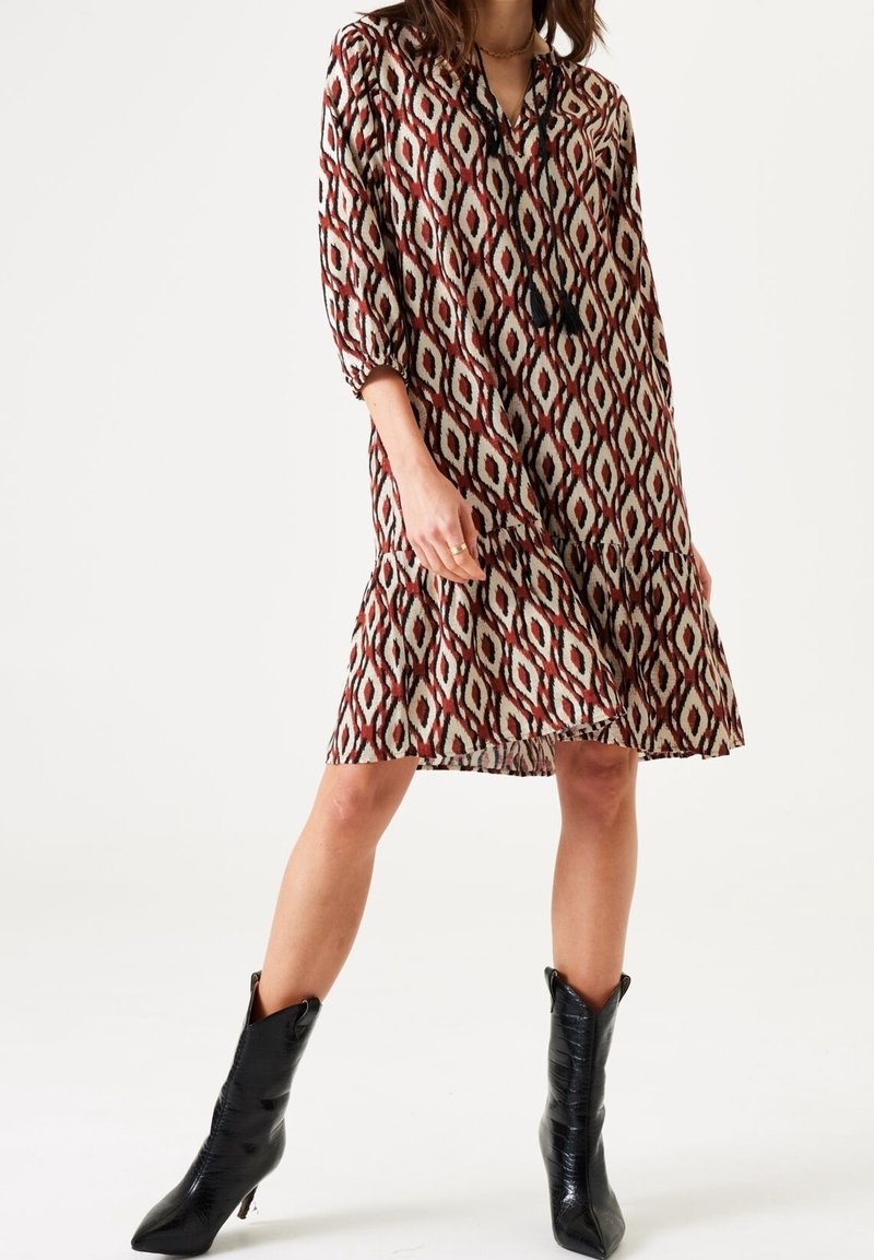 Femme portant une robe à motifs jusqu'aux genoux avec des manches trois-quarts et des bottes noires à bout pointu mi-mollet, debout les jambes écartées.