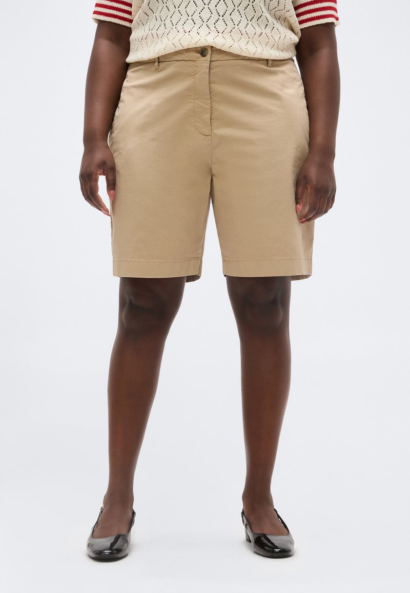 Άτομο που φοράει μπεζ σορτς μέχρι το γόνατο, μπλούζα με κοντά μανίκια και σχέδιο, και μαύρα παπούτσια με χαμηλό τακούνι, στέκεται μπροστά σε λευκό φόντο.