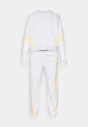 Szürke melegítő felső és nadrág szett, puha szövetből készült. Krém színű oldalsó panelek jellemzik, és diszkrét logó található a mellrészen. Sportos fazon.