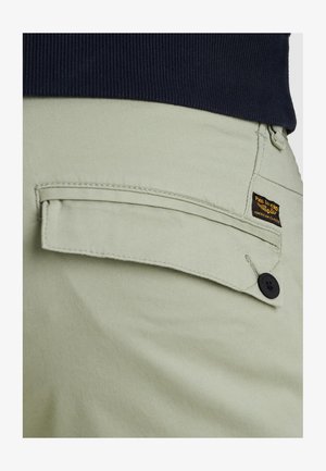 Helle grüne Hose mit Taschenklappe und schwarzem Knopf, kleines schwarz-gelbes PME Legend Markenetikett, kombiniert mit einem dunkel gerippten Oberteil.