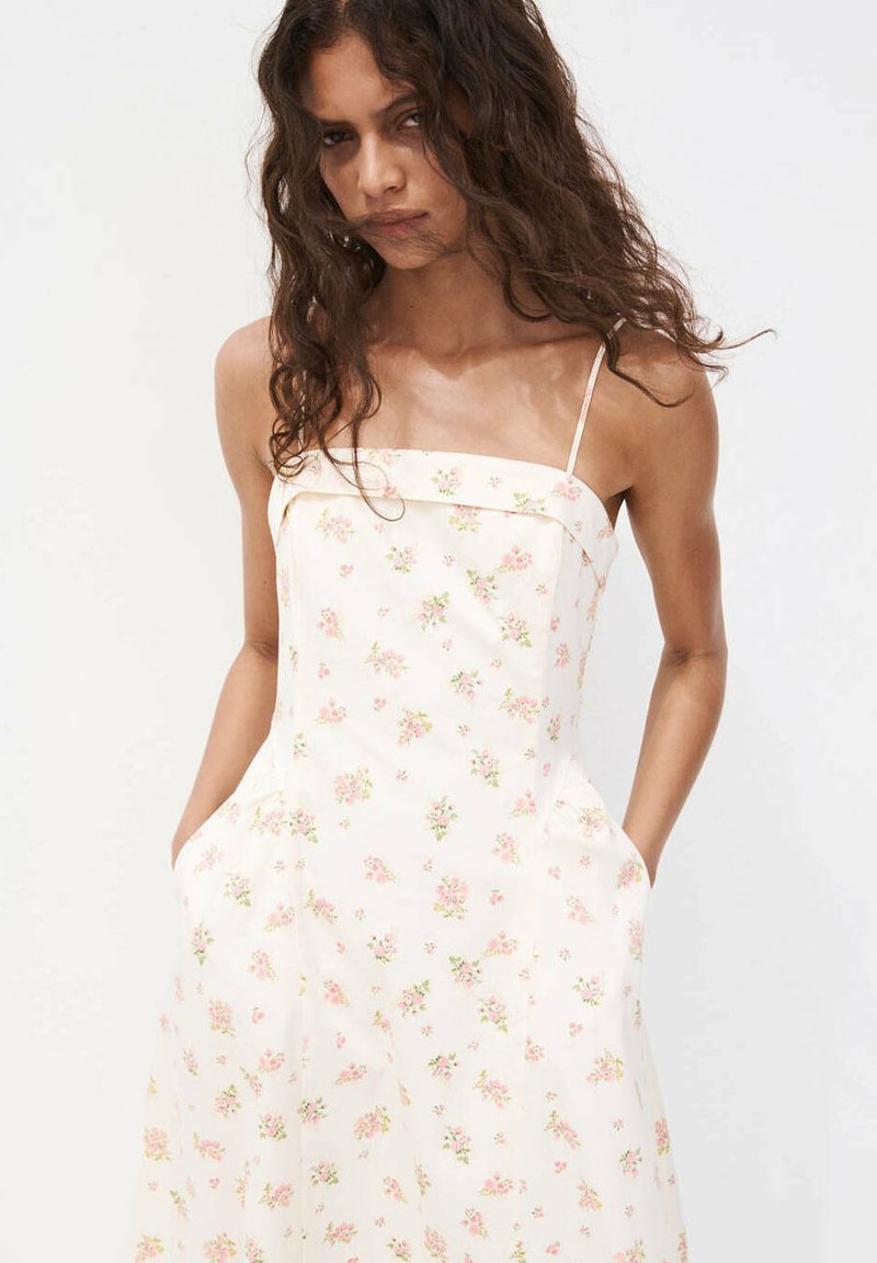 Vrouw met lang krullend haar die een witte mouwloze jurk draagt met dunne bandjes en kleine roze bloemmotieven, handen in de zakken.