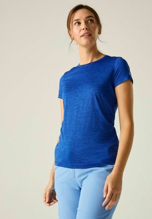 Joven mujer con una camiseta azul de manga corta y pantalones azul claro, de pie y mirando ligeramente hacia arriba y hacia un lado.