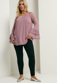 Blusa lunga a maniche con volant e tessuto testurizzato in rosa pallido, abbinata a leggings neri e scarpe a punta argentate.