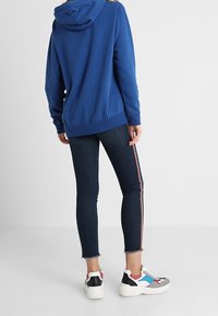 Blå hoodie med huva, kombinerad med mörka jeans med röda och vita sidostråkar. Sneakers har grå, rosa och blå accenter.
