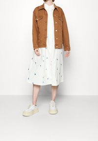 Veste en denim marron sur une robe blanche avec broderies florales bleues, associée à des baskets compensées blanches. Textures lisses, design décontracté.