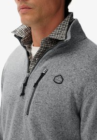 Szary polarowy sweter z półzamkiem, czarnymi elementami i tkaną koszulą w kratę pod spodem. Posiada naszywkę z logo na klatce piersiowej.