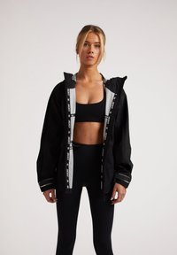 Svart vindjacka med grå innertyg, kontrasterande remmar och reflektiva detaljer. Buren över en svart träningslinne och leggings.