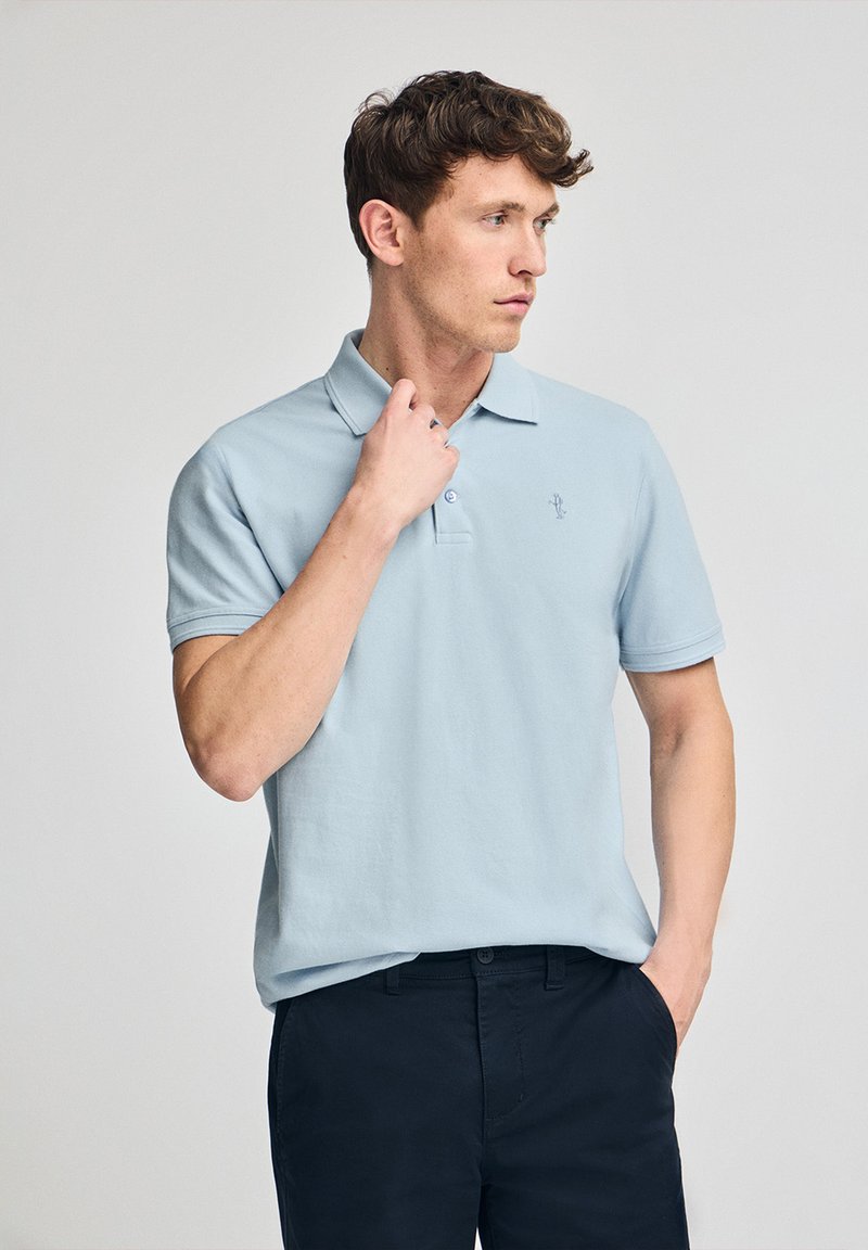 Camisa polo de color azul claro con cuello, mangas cortas y un logo bordado en el pecho. Se combina con pantalones oscuros. Textura de algodón suave.