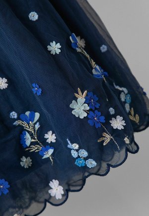 Granatowa tkanina tiulowa ozdobiona haftowanymi kwiatami w odcieniach niebieskiego, białego i złotego, z detalem falistego brzegu.