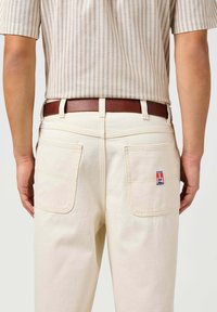 Beżowe spodnie z płótna z dwiema tylnymi kieszeniami, wyśrodkowaną łatą z logo oraz brązowym skórzanym paskiem; widoczna powyżej prążkowana bluzka z krótkim rękawem.