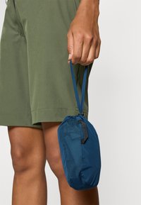 Blå reiseveske med nettdesign og snor-lukking, kompakt holdt i en hånd ved siden av olivengrønne shorts.