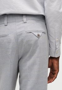 Άνδρας που φοράει ανοιχτό γκρι παντελόνι επίσημο με κουμπωτές τσέπες πίσω και εμφανές μανίκι ανοιχτογκρι πουκάμισου.