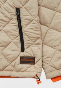 Veste matelassée beige dotée d'une fermeture éclair noire, de touches orange à l'ourlet, et d'une étiquette affichant "Updated Garments" avec une finition texturée.