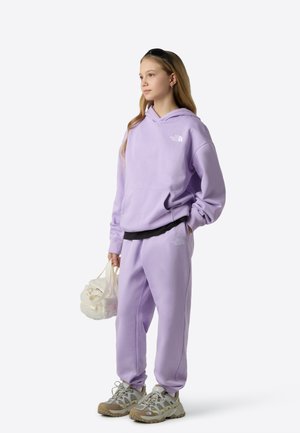 Sweat à capuche violet clair et joggings assortis, avec une poche avant et un logo. Associés à des baskets grises et un sac translucide.