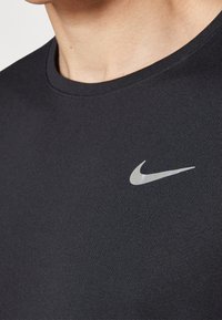 Camiseta negra de Nike con cuello redondo, mangas cortas y un logo de swoosh plateado reflectante en el lado izquierdo del pecho. Tejido suave y texturizado.