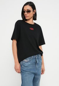 Μαύρο βαμβακερό t-shirt με κοντά μανίκια και κόκκινη λεπτομέρεια λογότυπου στο στήθος, συνδυασμένο με μπλε τζιν παντελόνι με διακοσμητικές γαλάζιες λεπτομέρειες.