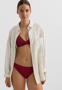 Lanena srajca v svetlo bež barvi z enim prsni žepom, nosena preko temno rdečega bikinija z teksturiranim vzorcem. Gladek, zračen material.