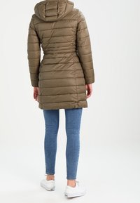 Abrigo de plumas verde oliva con capucha, con textura acolchada horizontal y diseño de cintura ajustada, combinado con vaqueros azules y zapatillas blancas.