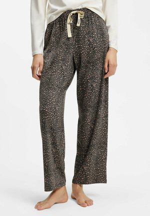 Femme portant un pantalon large imprimé léopard avec un cordon crème, debout pieds nus sur fond blanc, mains détendues le long du corps.