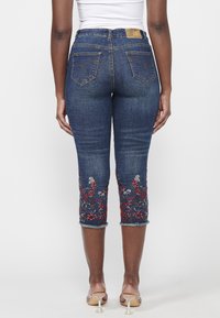 Dark blue denim capri jeans with floral embroidery in red and silver at the hem, featuring a slight fray and back pockets with decorative stitching.