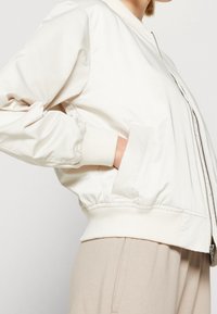 Světlobéžová bomber bunda se zipem vpředu, žebrovanými manžetami a lemem, s dvěma bočními kapsami a hladkou, lesklou texturou.