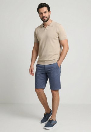 Mann steht mit einer Hand in der Tasche, trägt ein beiges Kurzarm-Poloshirt, blaue knielange Shorts und blaue Freizeitsneaker.