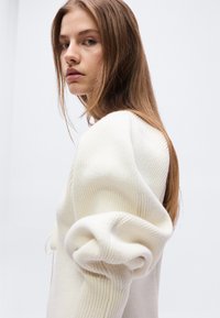 Νεαρή γυναίκα με μακριά, ανοιχτό καστανά μαλλιά, που φοράει λευκό ριμπ πουλόβερ με μεγάλα φουσκωτά μανίκια, κοιτώντας προς τα πίσω πάνω από τον ώμο της.