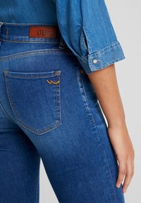Jeans de denim azul con un ajuste de cintura media, que presentan un parche de cuero marrón etiquetado como "LTB", y costuras sutiles en naranja en los bolsillos traseros.
