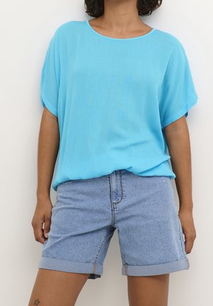 Person iført løs lyseblå kortærmet top og optrukne denimshorts, stående mod en ensfarvet hvid baggrund.