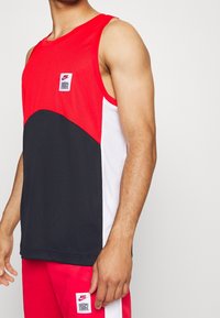 Débardeur sportif sans manches rouge et noir avec des accents blancs et un patch logo carré, doté d'un tissu texturé et d'un design sportif.