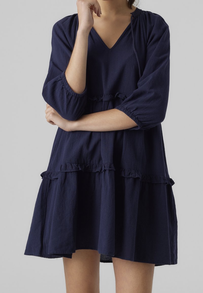 Kvinna klädd i en marinblå långärmad klänning med volangdetaljer, med armarna i kors och handen mot hakan framför en enfärgad bakgrund.