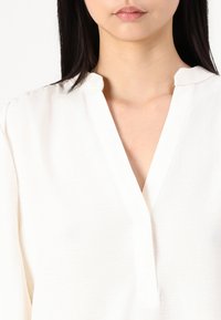 Hvit bluse med spiss krage og halv-bluss. Stoffet ser mykt og lett ut, med subtile teksturer og sømdetaljer.