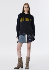 Pull en tricot bleu marine avec "INFINITE" en fil doré, associé à une jupe en denim avec fermeture éclair et graphismes, complété par des bottes en suède beige.