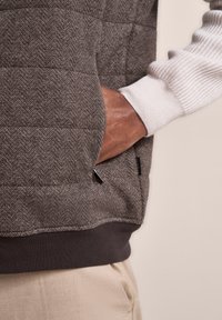 Gepolsterte Weste mit Fischgrätmuster in Dunkelgrau, ausgestattet mit einer Kängurutasche und gerippten weißen Bündchen, umrandet von einem dunklen Taillenbund.