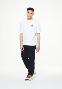 Camiseta blanca de algodón con mangas cortas y un logo negro en el pecho, combinada con pantalones negros y zapatillas de deporte con cordones multicolor.