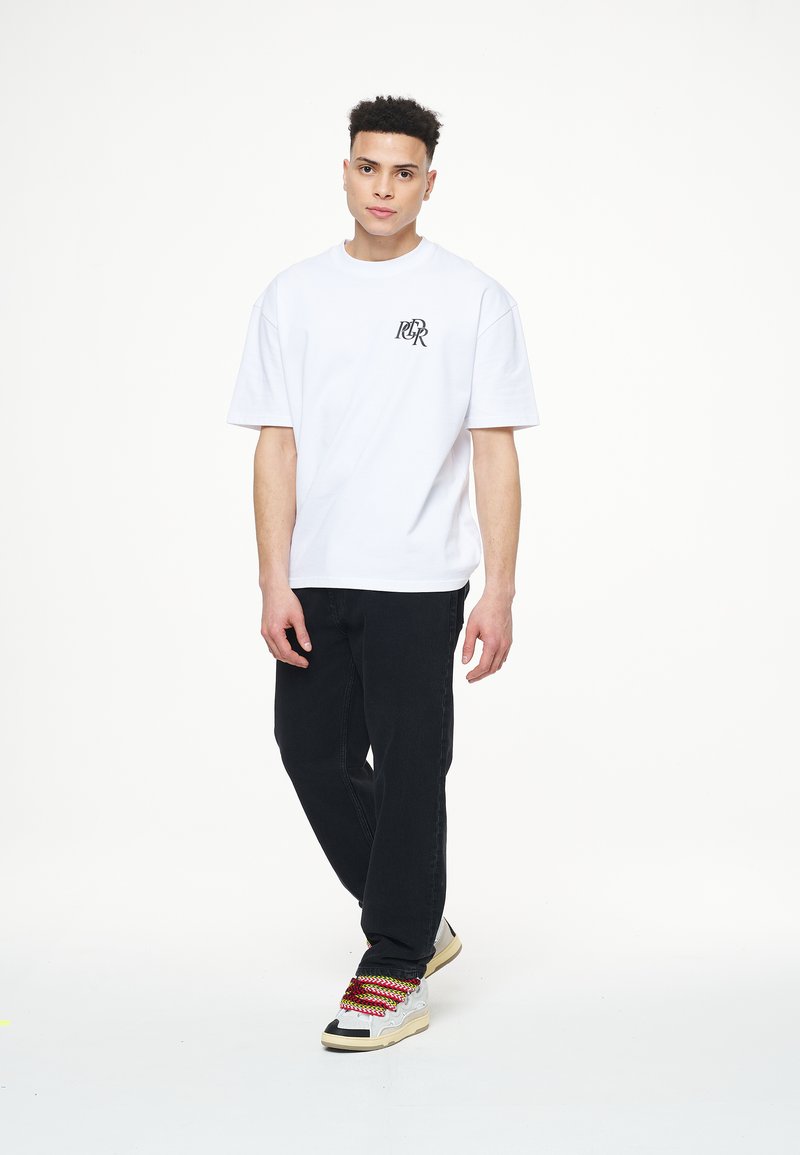 Camiseta blanca de algodón con mangas cortas y un logo negro en el pecho, combinada con pantalones negros y zapatillas de deporte con cordones multicolor.