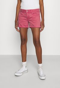 Short en denim rose avec fermeture éclair et bouton, revers roulés et poches avant, associé à des baskets montantes blanches.