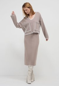 Ensemble en tricot de couleur marron clair avec un col en V, deux poches avant et des bords blancs, associé à une jupe longue assortie et des bottes crème.