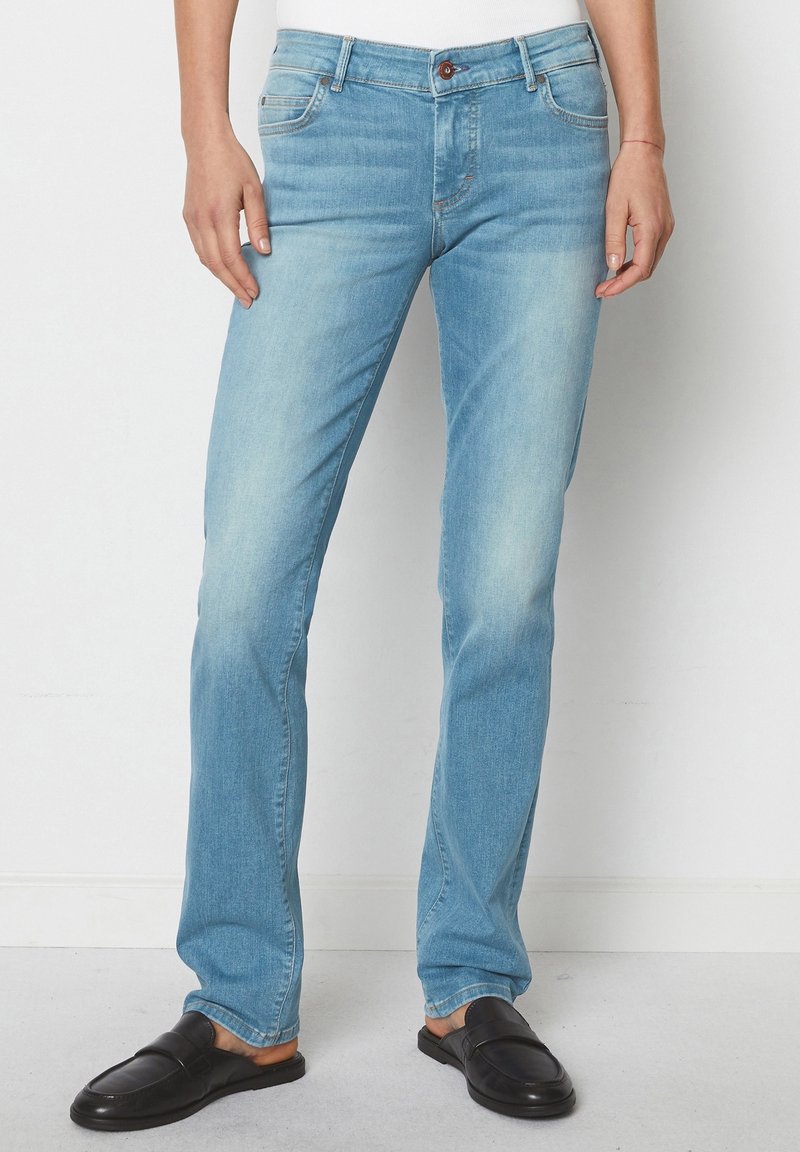 Personne portant un jean bleu clair à coupe droite et des mocassins noirs sans lacets, debout devant un fond blanc uni.