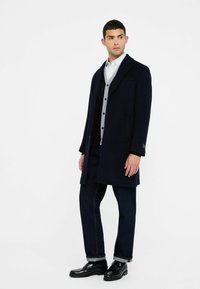 Man in een marineblauwe overjas, lichtgrijze geknoopte cardigan, wit shirt, donkere spijkerbroek met omslagen en zwarte leren loafers, staand tegen een witte achtergrond.