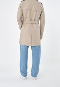Trench coat beige con dettaglio in vita sul retro, maniche lunghe e spalline sulle spalle, abbinato a jeans a gamba larga blu chiaro e sneakers bianche.