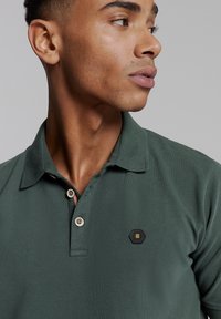 Donkergroene polo met een gestructureerde stof, drie knopen en een klein hexagonaal logo op de linkerkant van de borst. Classiek ontwerp van de kraag.