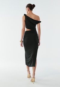 Vrouw met haar in een knot, gekleed in een zwarte asymmetrische jurk met een open rug, gouden armband, ronde oorbellen en gouden sandalen met bandjes, die wegloopt.