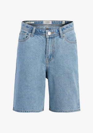 Garçon debout regardant vers la droite, portant une chemise blanche à manches courtes avec motif, un short en jean, des chaussettes rayées et des baskets blanches.