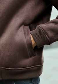Hanorac din fleece maro, cu buzunar lateral, mansete striate și o textură netedă, având o mână vizibilă așezată în buzunar.