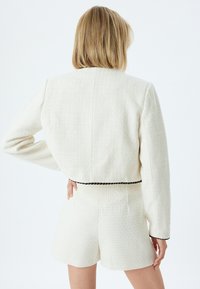 Giacca da suit in tessuto bianco con una vestibilità cropped, caratterizzata da rifiniture nere in vita e maniche lunghe classiche, abbinata a pantaloni corti coordinati.